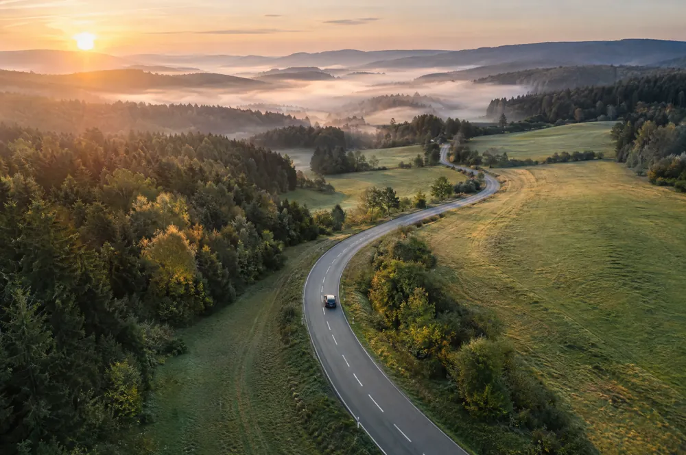 Auto auf einer kurvigen Landstraße bei Sonnenaufgang aus der Luft